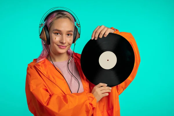 Hezká dívka s růžovými vlasy si užívá poslouchání hudby. Hipsterová žena s mimořádným vzhledem v oranžovém plášti drží vinylovou desku na modrém pozadí. — Stock fotografie
