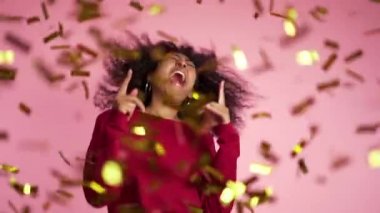 Afro saçlı, güzel Afro-Amerikalı bir kadın. Stüdyoda pembe arka planda konfeti yağmuru altında gülüyor ve dans ediyor. yavaş çekim