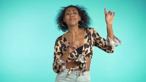 Прекрасна афро-американська жінка з африканським волоссям, що веселиться посміхаючись і танцюючи в студії на синьому фоні. повільний рух — стокове відео