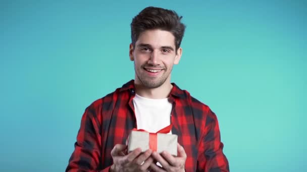 Handsome man holding christmas gift and gives it by hands it to camera. He is happy, smiling. Guy on blue background. Positive holiday footage. — Stock Video