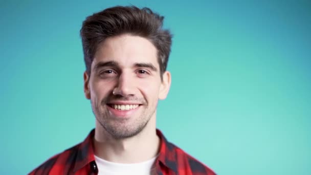 Hombre europeo guapo con peinado de moda en camisa a cuadros roja sobre fondo de estudio azul. Copiar espacio. Chico alegre sonriendo y mirando a la cámara . — Vídeo de stock