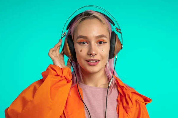 Το όμορφο κορίτσι με τα ροζ μαλλιά απολαμβάνει να ακούει μουσική. Hipster γυναίκα με εξαιρετική εμφάνιση σε πορτοκαλί αδιάβροχο σε μπλε φόντο. — Φωτογραφία Αρχείου