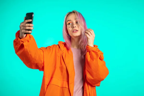 Menina com cabelo rosa e aparência extraordinária fazer selfie no fundo azul. Usando a tecnologia moderna smartphone, redes sociais — Fotografia de Stock