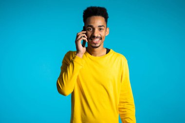 Sarı elbiseli gülümseyen Afrikalı Amerikalı adam mavi arka planda cep telefonuyla konuşuyor. Akıllı telefon kullanan yakışıklı bir adam. Teknolojili adam.