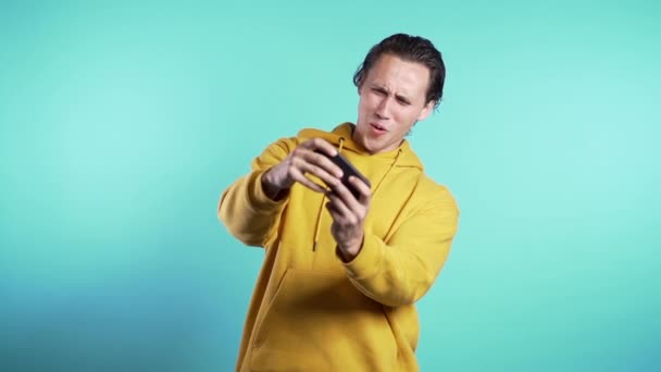 Hombre guapo jugando en el teléfono inteligente en la pared del estudio azul. Uso de la tecnología moderna - aplicaciones, redes sociales. Movimiento lento — Vídeo de stock