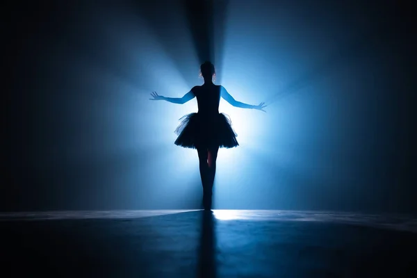 Fiatal gyönyörű balerina a füstszínpadon táncol modern balett. Végrehajtja sima mozdulatok kezével szemben reflektorfény háttér. Nő fekete tütü jelmezben a helyszínen. — Stock Fotó