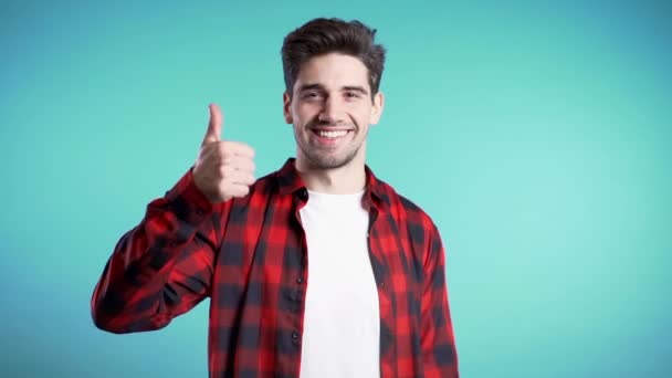 El joven positivo sonríe a la cámara. Hipster tipo mostrando el pulgar hacia arriba signo sobre fondo azul. Ganador. Éxito. Lenguaje corporal . — Vídeo de stock