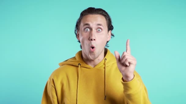 Portrét mladého přemýšlejícího muže ve žlutém, který má představu o momentu ukazujícím prstem nahoru na pozadí modrého studia. Usmívající se šťastný student chlap ukazující eureka gesto. — Stock video