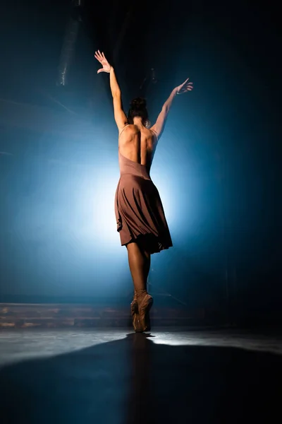 Gracieuze ballerina in beige jurk dansende elementen van klassiek of modern ballet in het donker met achtergrondverlichting. Rook op zwarte achtergrond. Kunstconcept. — Stockfoto