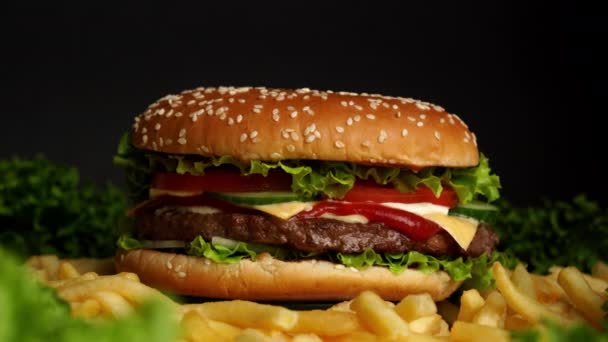 Hambúrguer de carne suculenta com costeleta, cebola, legumes, queijo derretido, alface, molho e sementes de gergelim cobertas. Hambúrguer isolado gira sobre fundo escuro, vista close-up — Vídeo de Stock