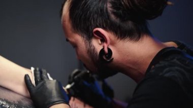 Dövmeci, müşterisi için makineyle dövme yapıyor. Parlorda dövme yaptırma işlemi. Vücut dövme işlemi. Rastalı bir adam lambanın altında kadın vücudu üzerinde çalışıyor..