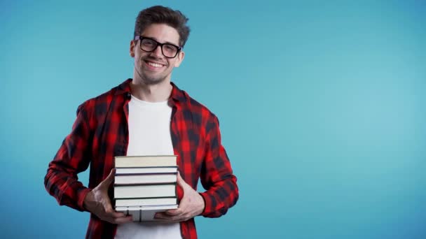 Der europäische Student im rot karierten Hemd auf blauem Hintergrund hält im Atelier einen Stapel Universitätsbücher aus der Bibliothek. Kopierraum. Mann lächelt, er ist glücklich, seinen Abschluss zu machen. — Stockvideo