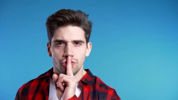 Ένας όμορφος Ευρωπαίος που κρατάει το δάχτυλο στα χείλη του πάνω από μπλε φόντο. Χειρονομία σιωπής, μυστικότητας, σιωπής. Αντιγραφή χώρου — Αρχείο Βίντεο