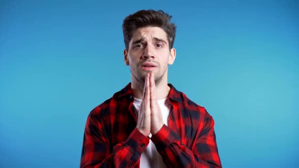 Hombre guapo con camisa roja rezando sobre fondo azul. Un tipo de raza europea rogando a alguien. 4k. Concepto de religión . — Vídeo de stock