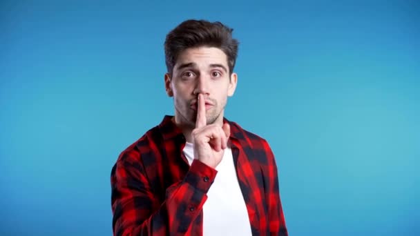 Ένας όμορφος Ευρωπαίος που κρατάει το δάχτυλο στα χείλη του πάνω από μπλε φόντο. Χειρονομία της σιωπής, μυστικό, σιωπή — Αρχείο Βίντεο