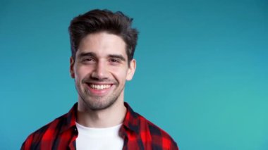 Mavi stüdyo arka planında kırmızı ekose gömlekli yakışıklı Avrupalı bir adam. Gülümseyen ve kameraya bakan neşeli adam. 4k