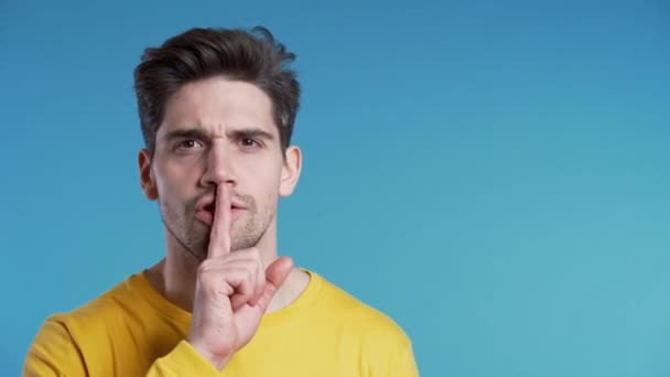 Avrupalı yakışıklı bir adam mavi arka planda parmaklarını tutuyor. Şşş, sır, sessizlik jesti. Boşluğu kopyala — Stok video