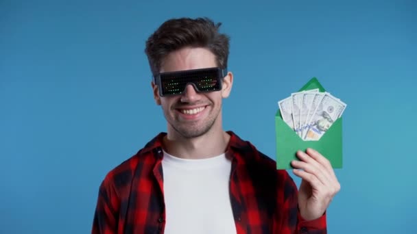 Szczęśliwy Europejczyk w świecących okularach znaków dolara pokazujących pieniądze w kopercie - dolary amerykańskie banknoty na niebieskiej ścianie. Symbol sukcesu, zysku, zwycięstwa — Wideo stockowe