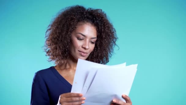 Une jolie patronne afro-américaine vérifie les documents, les factures d'électricité. Femme d'affaires sérieuse debout sur fond bleu. Elle est satisfaite du travail du personnel. 4k — Video