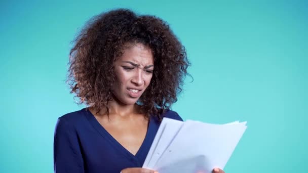 La femme africaine sérieuse n'est pas satisfaite des papiers, des factures, des documents ou des rapports sur fond bleu. Jeune employée de bureau isolée en studio. 4k — Video