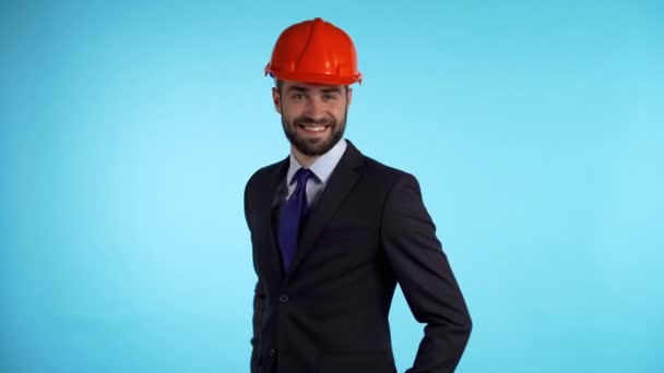 Empresário jovem engenheiro positivo no capacete de construção sorri para a câmera. Homem mostrando polegares para cima sinal sobre fundo azul. Vencedor. Sucesso. Linguagem corporal. 4k — Vídeo de Stock