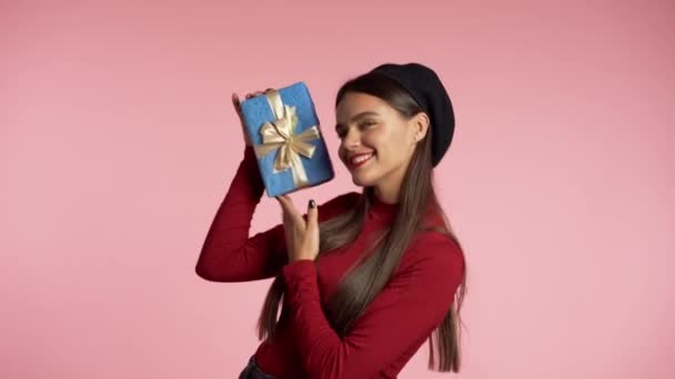 Радісна красива жінка з ідеальним макіяжем, що тримає синю подарункову коробку з бантом на рожевому тлі стін. Модна дівчина посміхається, вона рада бути присутньою . — стокове відео