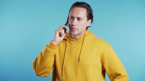 Egy sárga ruhás fiatalember beszél a mobilján, kék háttérrel. A trendi srác szokott beszélgetni. Okostelefon, technológiai koncepció. — Stock videók
