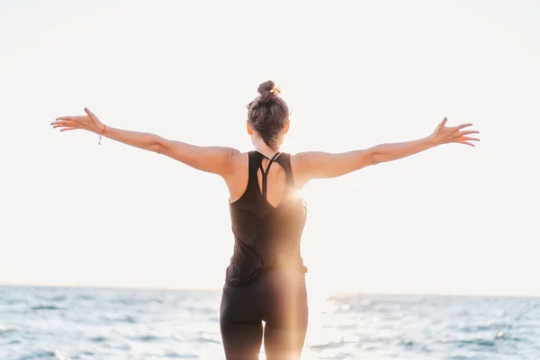 Vékony nő fekete testruhában jógázik a tenger vagy az óceán közelében napfelkeltekor. Rugalmasság, nyújtás, fitness, egészséges életmód. — Stock Fotó