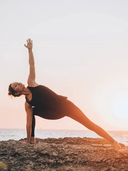 Donna magra in body nero che pratica yoga vicino al mare o all'oceano durante la luce dell'alba. Flessibilità, stretching, fitness, stile di vita sano . — Foto Stock
