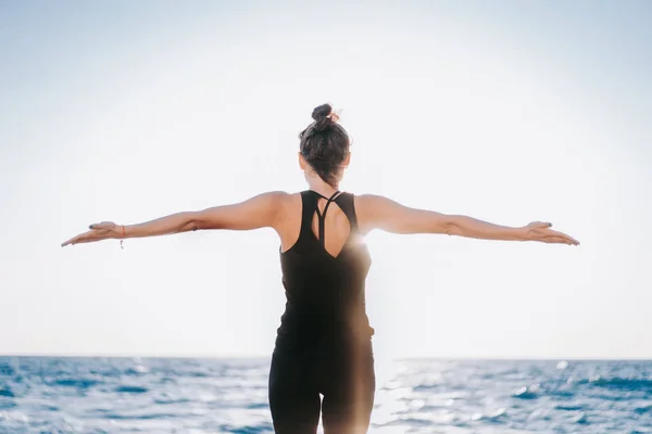 Vékony nő fekete testruhában jógázik a tenger vagy az óceán közelében napfelkeltekor. Rugalmasság, nyújtás, fitness, egészséges életmód. — Stock Fotó