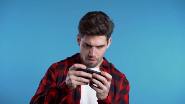 Schöner Mann spielt Spiel auf Smartphone an blauer Studiowand. — Stockvideo
