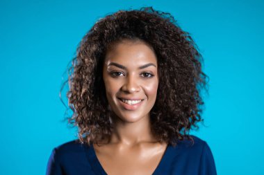 Afro saçlı, iş kıyafetleri içinde, mavi duvar arkasından kameraya gülümseyen çekici Afrikalı Amerikalı kadın. Şirin melez kızlar portresi.