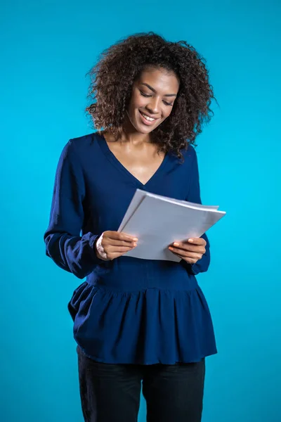 Una jefa afroamericana chequea documentos, facturas de servicios públicos. Mujer de negocios seria de pie sobre fondo azul. Ella está satisfecha con el trabajo del personal . —  Fotos de Stock