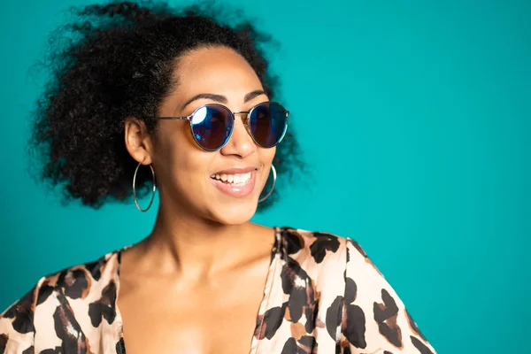 Hübsche afrikanische Frau mit Afro-Haaren im Leopardenoberteil und Sonnenbrille, die vor blauem Wandhintergrund in die Kamera lächelt. Niedliche gemischte Rasse Mädchen Porträt. — Stockfoto