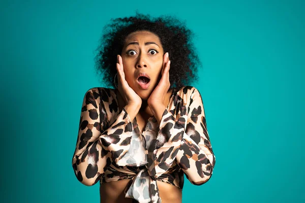 Frightened african american woman with curly hair in leopard shirt afraid of something and looks into the camera with big eyes full of horror over blue wall background.