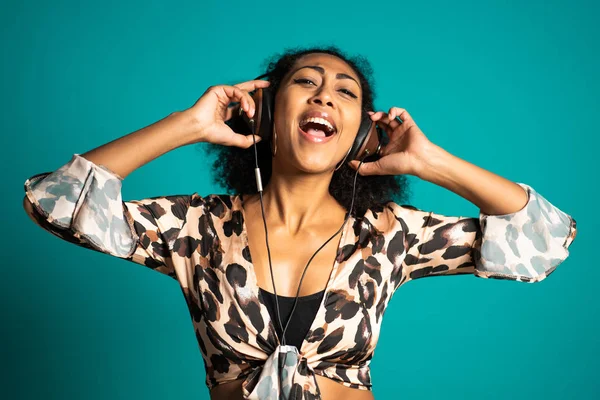 Piękna Afroamerykanka z afro włosy i duże słuchawki zabawy uśmiecha się i tańczy w studio na niebieskim tle. — Zdjęcie stockowe