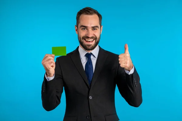 Успішна людина в діловому костюмі, що показує необмежену зелену кредитну картку на синьому фоні студії. Професійний чоловічий менеджер . — стокове фото