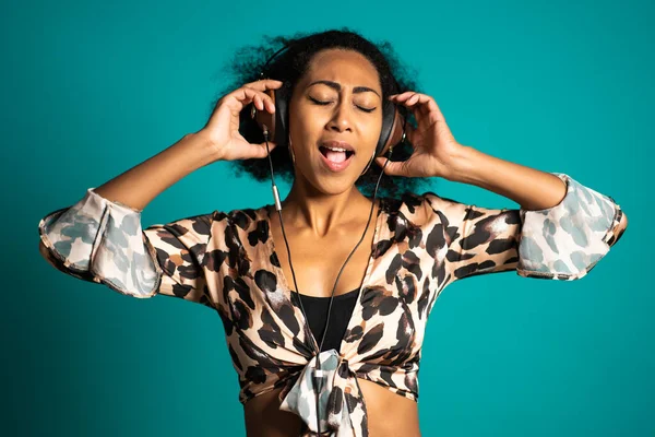 Beautiful african american woman with afro hair and big headphones having fun smiling and dancing in studio against blue background.