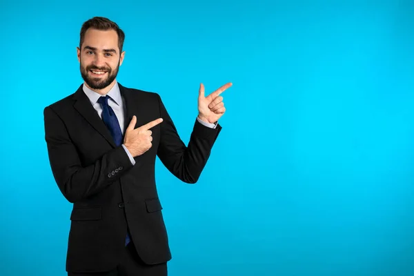 青い背景に孤立した何かを示す、企業のスーツのプレゼンテーションで幸せな笑顔のビジネスマン。男の肖像画、彼はコピースペースで彼の左側に腕を指している. — ストック写真