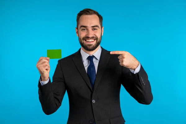 Homem de sucesso em terno de negócios mostrando cartão de crédito verde ilimitado em fundo estúdio azul. Profissional gerente masculino . — Fotografia de Stock