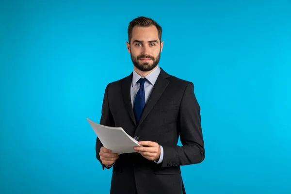 Jovem bonitão de casaco com documentos, contas, relatório ou contrato. Empresário isolado em fundo azul. Ele está satisfeito. . — Fotografia de Stock