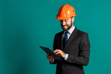 Takım elbiseli genç mühendis, inşaat projesiyle ilgili tablete notlar yazıyor. Rapor ver. İşadamı, inşaatta olduğu gibi mavi arka planda da izole edilmiş. Tatmin oldu..