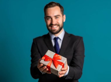 Takım elbiseli yakışıklı iş adamı hediye kutusunu verir ve kameraya verir. O mutlu, gülümsüyor. Mavi arka planda sakallı bir adam. Pozitif tatil çekimi.