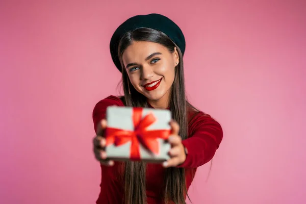 Pretty woman gives gift and hands it to the camera. She is happy, smiling. Girl on pink background. Positive holiday shot.
