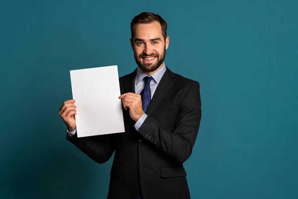Podnikatel drží bílý svislý plakát z papíru A4. Rozumím. Usmívající se pohledný muž v firemním obleku s kravatou na modrém pozadí. — Stock fotografie