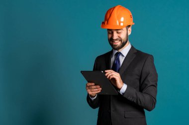 Takım elbiseli genç mühendis, inşaat projesiyle ilgili tablete notlar yazıyor. Rapor ver. İşadamı, inşaatta olduğu gibi mavi arka planda da izole edilmiş. Tatmin oldu..