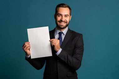 Elinde beyaz dikey a4 kağıt poster tutan bir iş adamı. Uzayı kopyala Mavi arka planda kravatlı, takım elbiseli, gülümseyen yakışıklı adam..