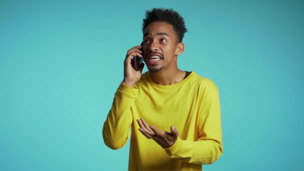 Angry african manin yellow wear screaming down, swears with somebody. Using mobile phone. Stressed and depressed man on blue background. — Stock Video