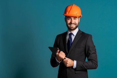 Takım elbiseli genç mühendis elinde inşaat projesiyle tablet tutuyor. İşadamı, inşaatta olduğu gibi mavi arka planda da izole edilmiş. Teknoloji, modern işgal kavramı.