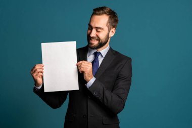 Elinde beyaz dikey a4 kağıt poster tutan bir iş adamı. Uzayı kopyala Mavi arka planda kravatlı, takım elbiseli, gülümseyen yakışıklı adam..